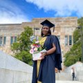 A young African woman stands in graduation cap and gown and holding flowers outside UQ's sandstone Great Court.